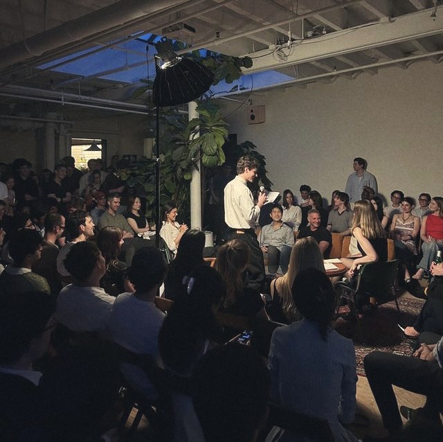 A packed loft audience seated on the floor watching a speaker with a microphone