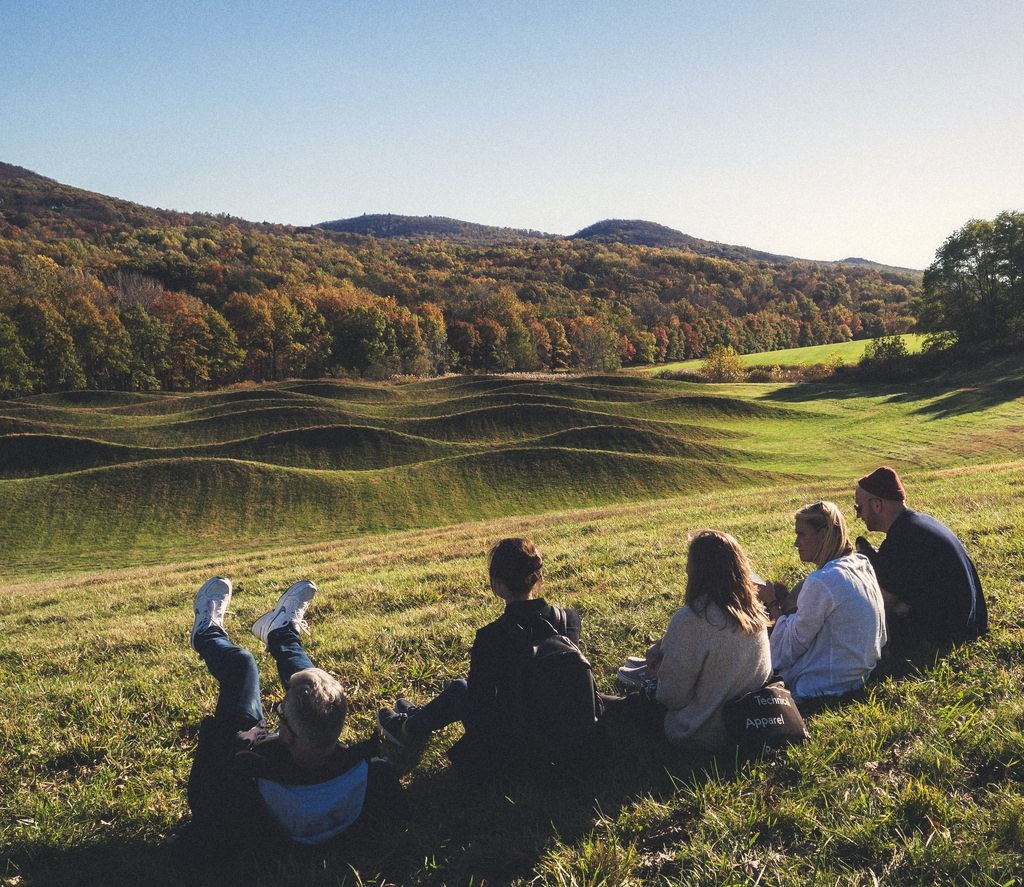 A group sitting on a grass hillside looking out at rolling fields and autumn woods