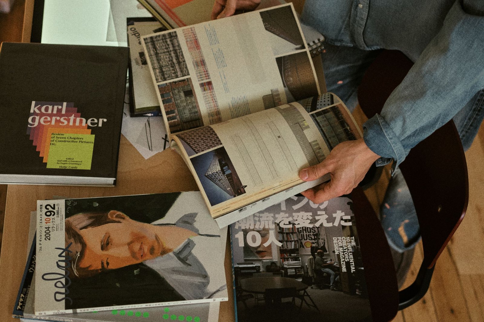 Hands leafing through an open design book on a table, next to Karl Gerstner&rsquo;s Review of Seven Chapters of Constructive Pictures, Etc. and stacked Japanese magazines