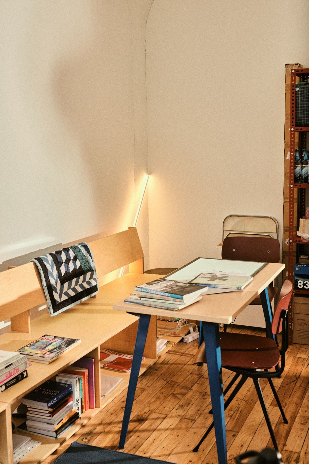 A sunlit corner with a plywood bench and shelves of design books, a quilt draped over the backrest, and a table with blue legs stacked with magazines