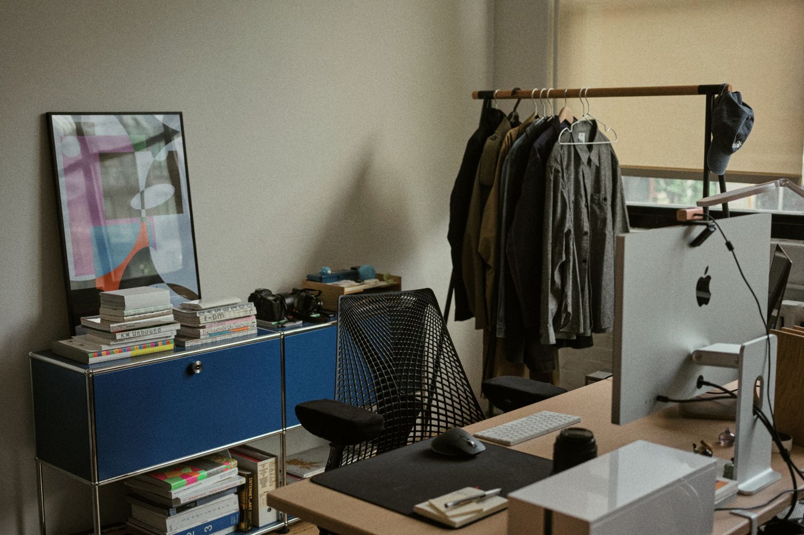 A desk with a large monitor, a mesh office chair, a blue USM cabinet stacked with design books, and a clothing rail by the window