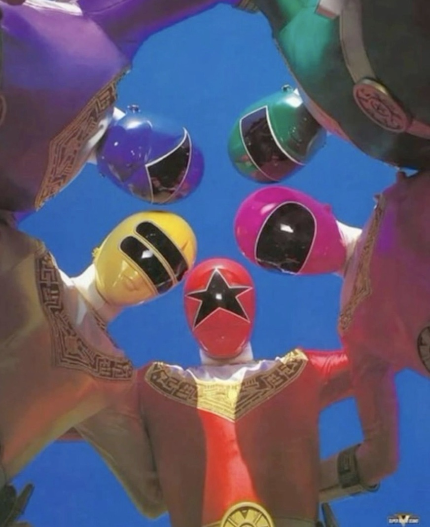 Five Power Rangers in colored helmets seen from below in a circle against a blue sky