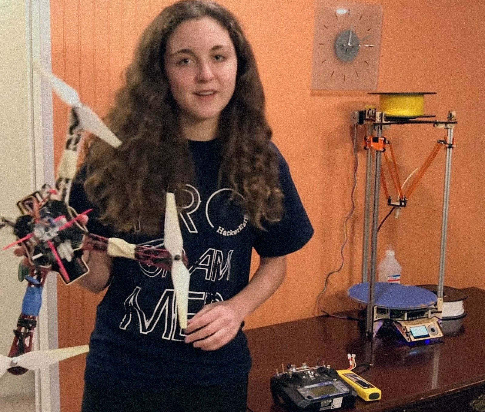 Young Liz holding a homemade drone, with a 3D printer and radio controller on the table behind her